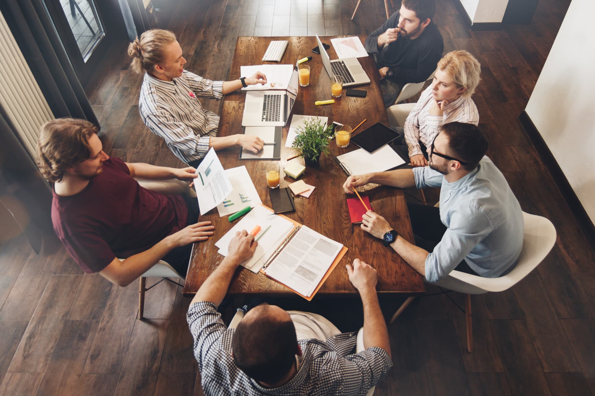 Six colleagues around a wooden table with laptops, printed charts, notebooks and highlighters, viewed from above during a working meeting.