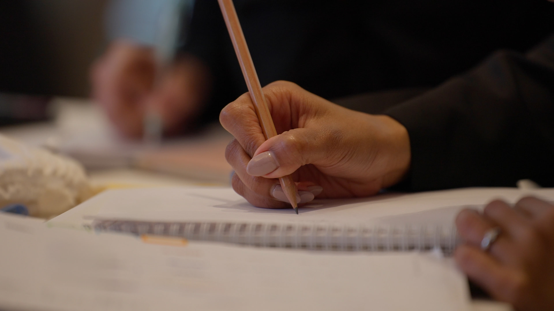 A close view of a hand writing in pencil on a page beside a spiral notebook, with another person writing in the background.