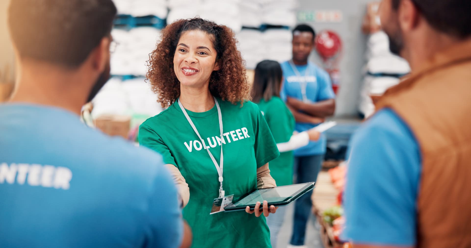 A volunteer in a green shirt holding a tablet and greeting a colleague inside a community food pantry, with other volunteers working behind her.