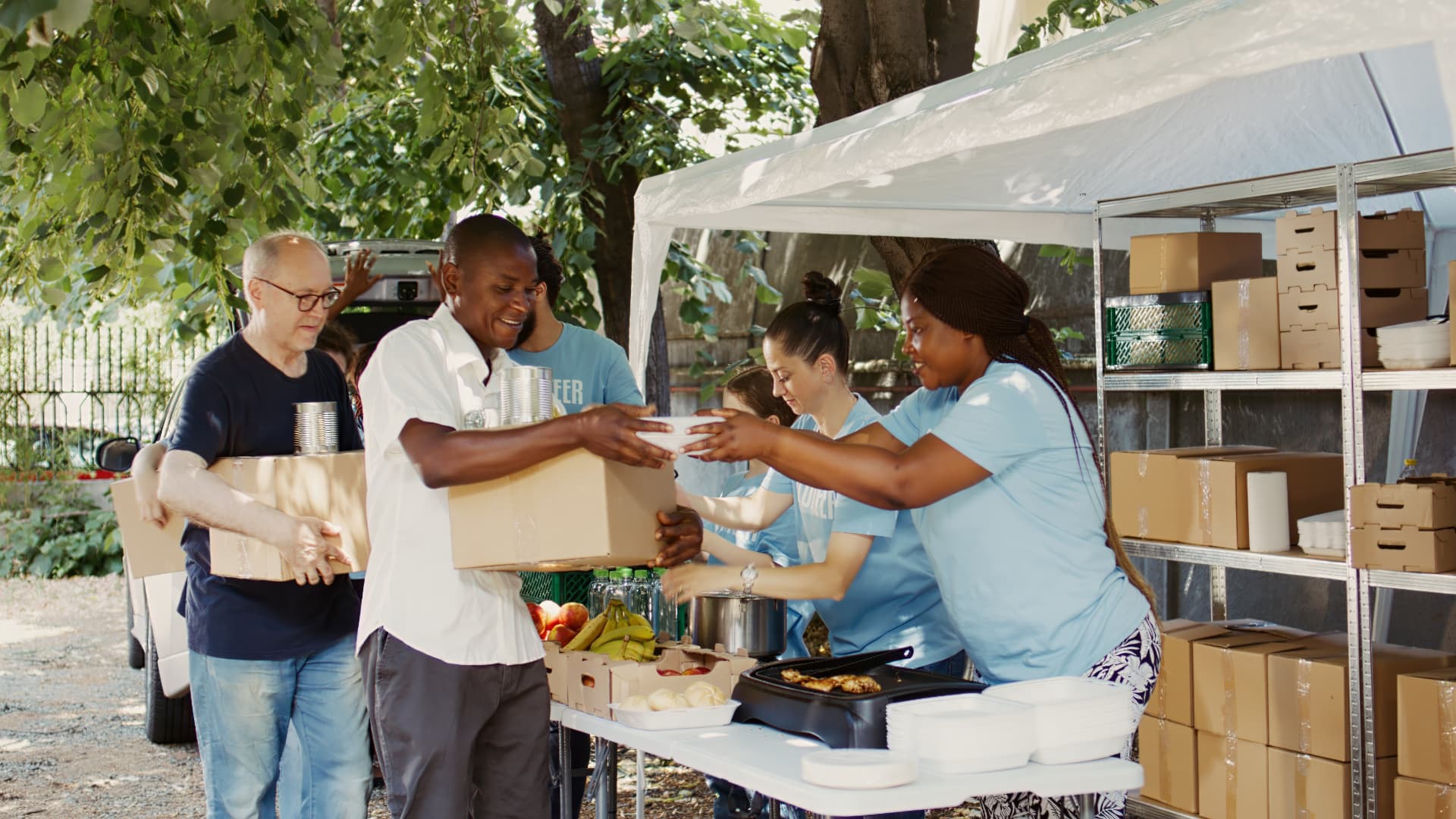 Volunteers under an outdoor canopy handing boxes and containers of food across a folding table, with shelves of supplies behind them.