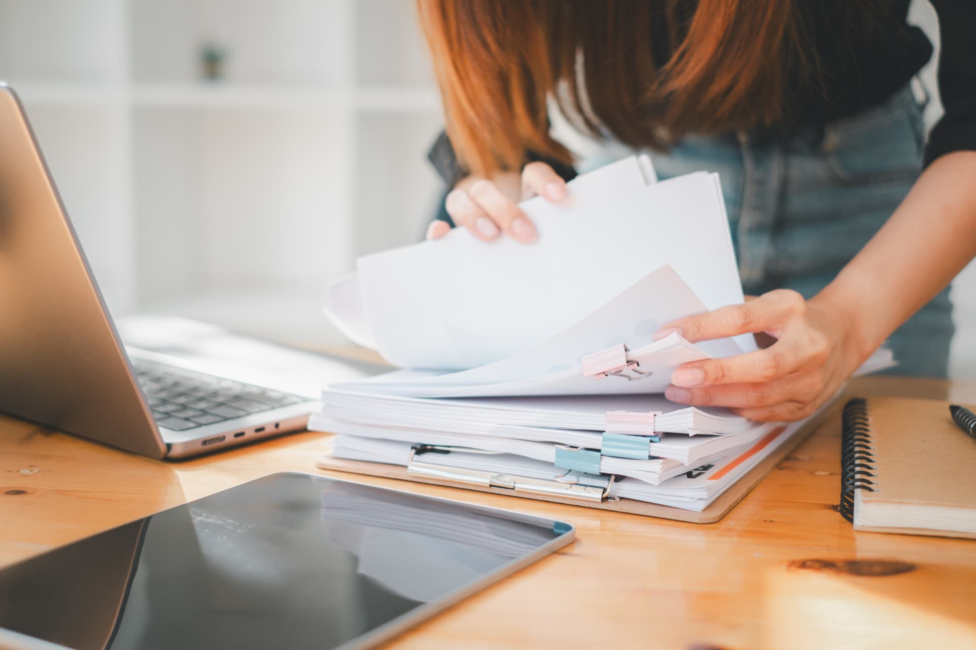 Hands sorting through a thick stack of clipped paperwork on a wooden desk beside an open laptop, a tablet and a notebook.